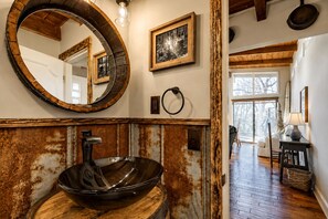 Un baño rústico con un espejo redondo grande, un lavabo de madera y un cuadro con marco de madera en la pared.