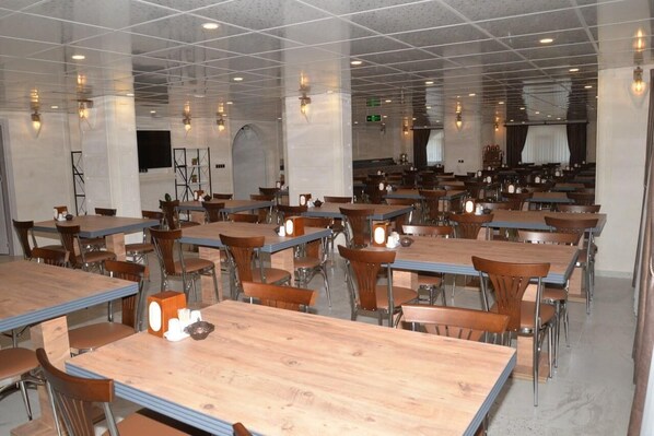 A spacious dining area with multiple tables and chairs, a high ceiling with grid-like patterns, and wall-mounted lighting fixtures.