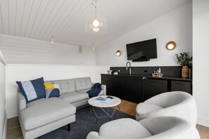 A modern living room with a sofa, armchairs, a coffee table, a flat-screen TV, and a kitchen area with a black countertop and cabinets.