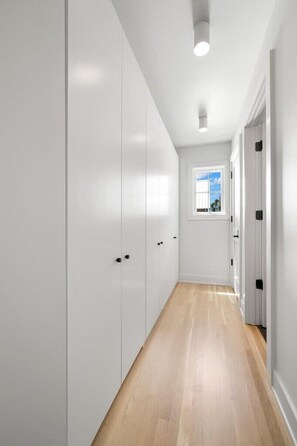 A narrow hallway with white walls and wooden flooring, leading to a window with a view of the sky.