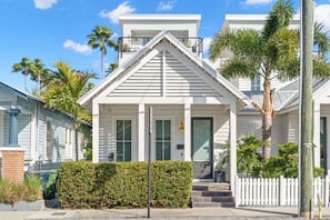A white two-story house with a front porch, a white picket fence, and palm trees in the background. - Xavier at Angels Quarter | Rooftop Terrace (Tampa)