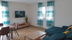 A living room with a wooden coffee table, a blue sofa, a TV mounted on the wall, and a dining area with a table and chairs. - Vacation apartments Hesselperle (Gerolfingen)