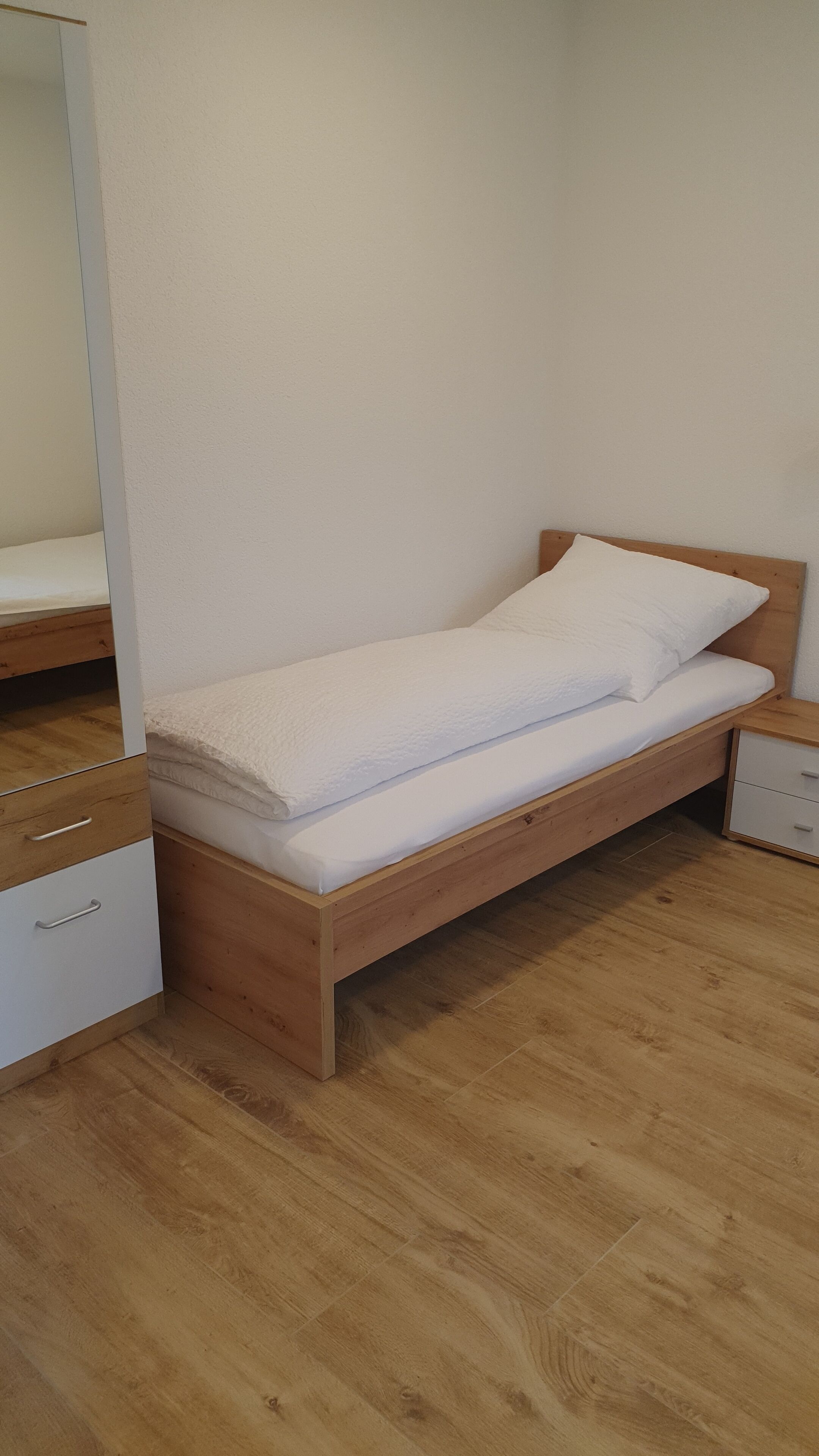 A single bed with a wooden frame and white bedding in a room with wooden flooring and white walls.