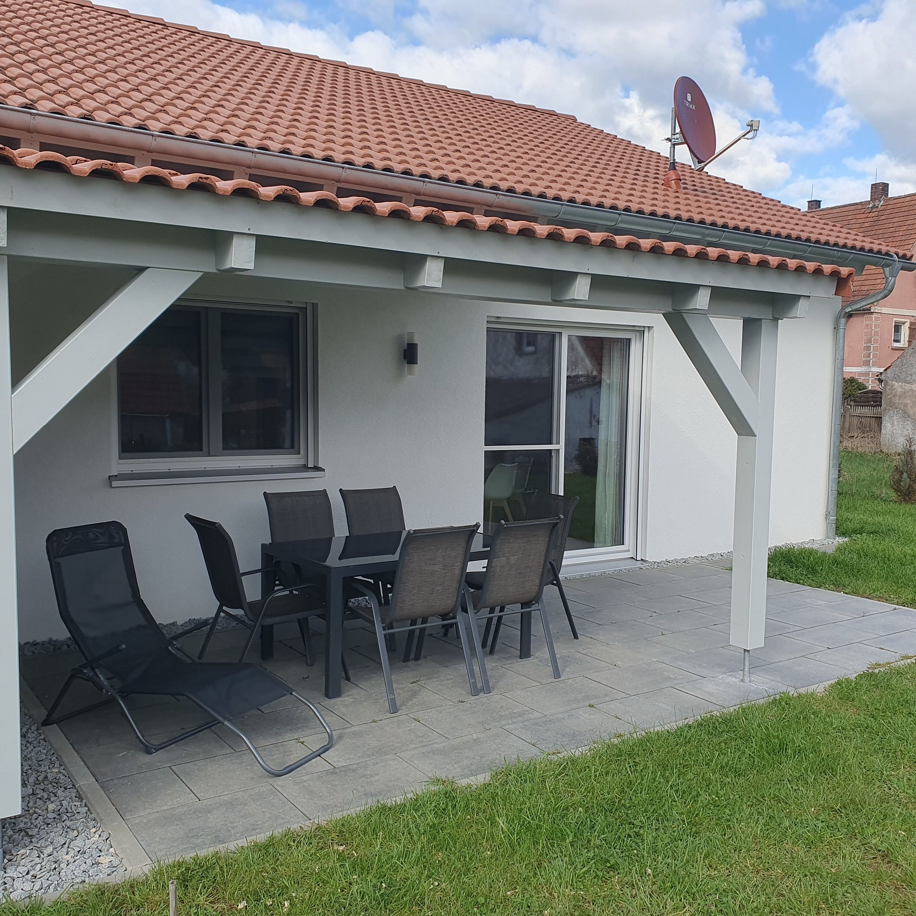 Ein kleines Haus mit einem Ziegeldach, einer Terrasse mit Tisch und Stühlen sowie einer Satellitenschüssel.