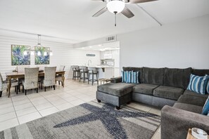 Un salon moderne avec un canapé d’angle gris, un coin repas avec des chaises en osier et une cuisine en arrière-plan.