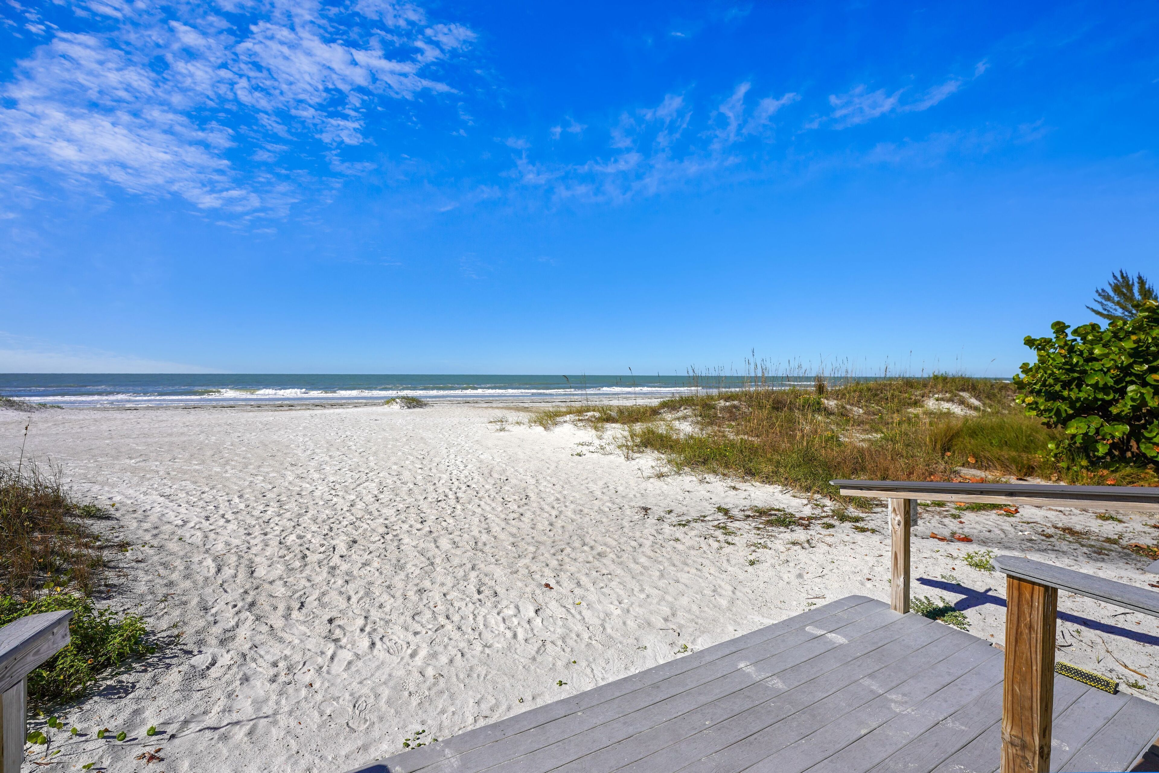 Une promenade en bois mène à une plage de sable fin, sous un ciel bleu et dégagé.