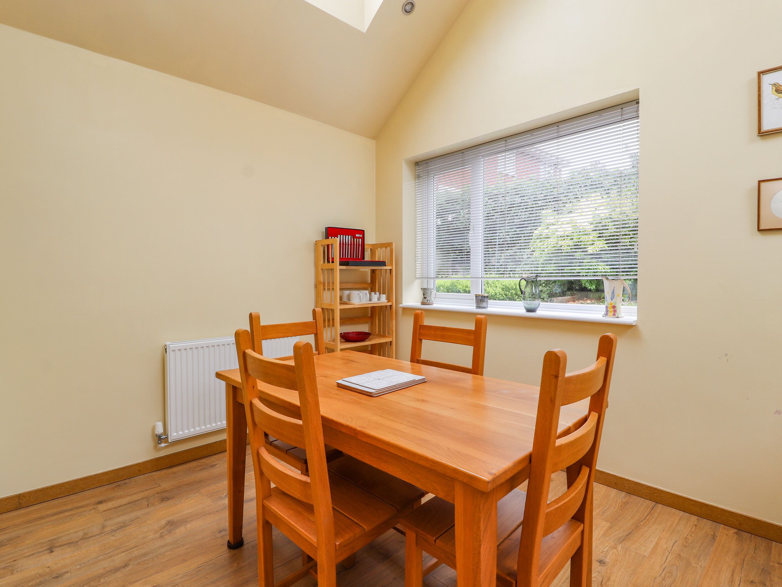 Un comedor con mesa y sillas de madera, un radiador, una estantería y una ventana con persianas.