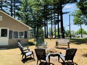 A house with a front yard featuring three Adirondack chairs, a wooden bench, and a small fire pit.