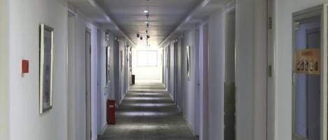 A long, narrow hotel corridor with doors on both sides, a fire alarm on the left, and a poster on the right wall.