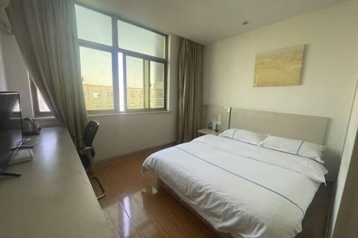 A hotel room with a bed, a desk, a chair, a window with curtains, and a painting on the wall.