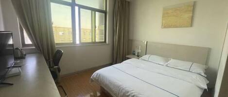 A hotel room with a bed, a desk, a chair, a window with curtains, and a painting on the wall.