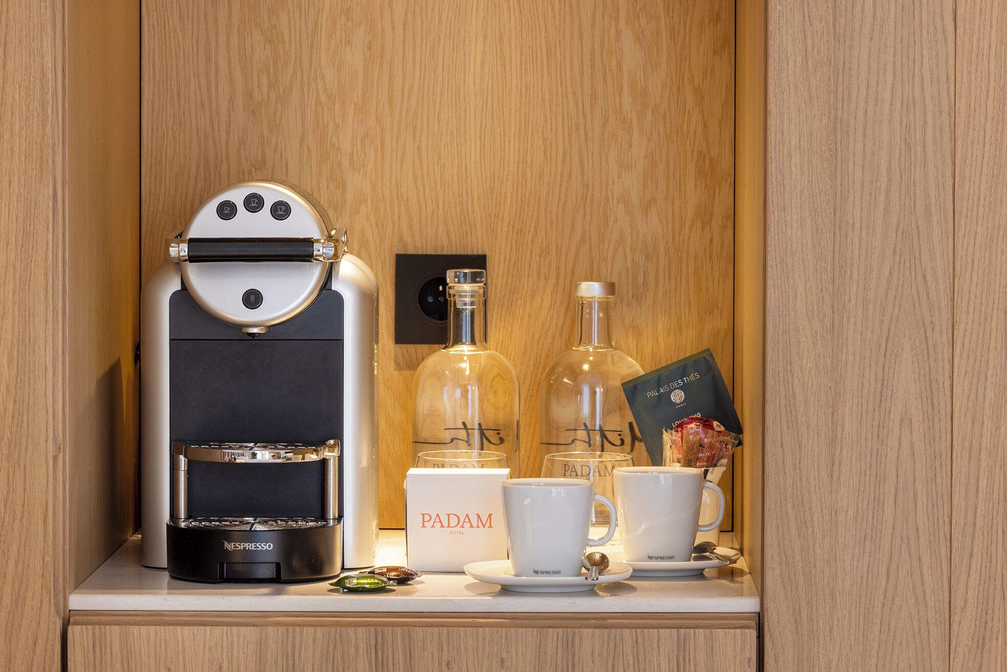 A minibar with a Nespresso coffee machine, two bottles of wine, a card, and two white cups with a Nespresso logo.