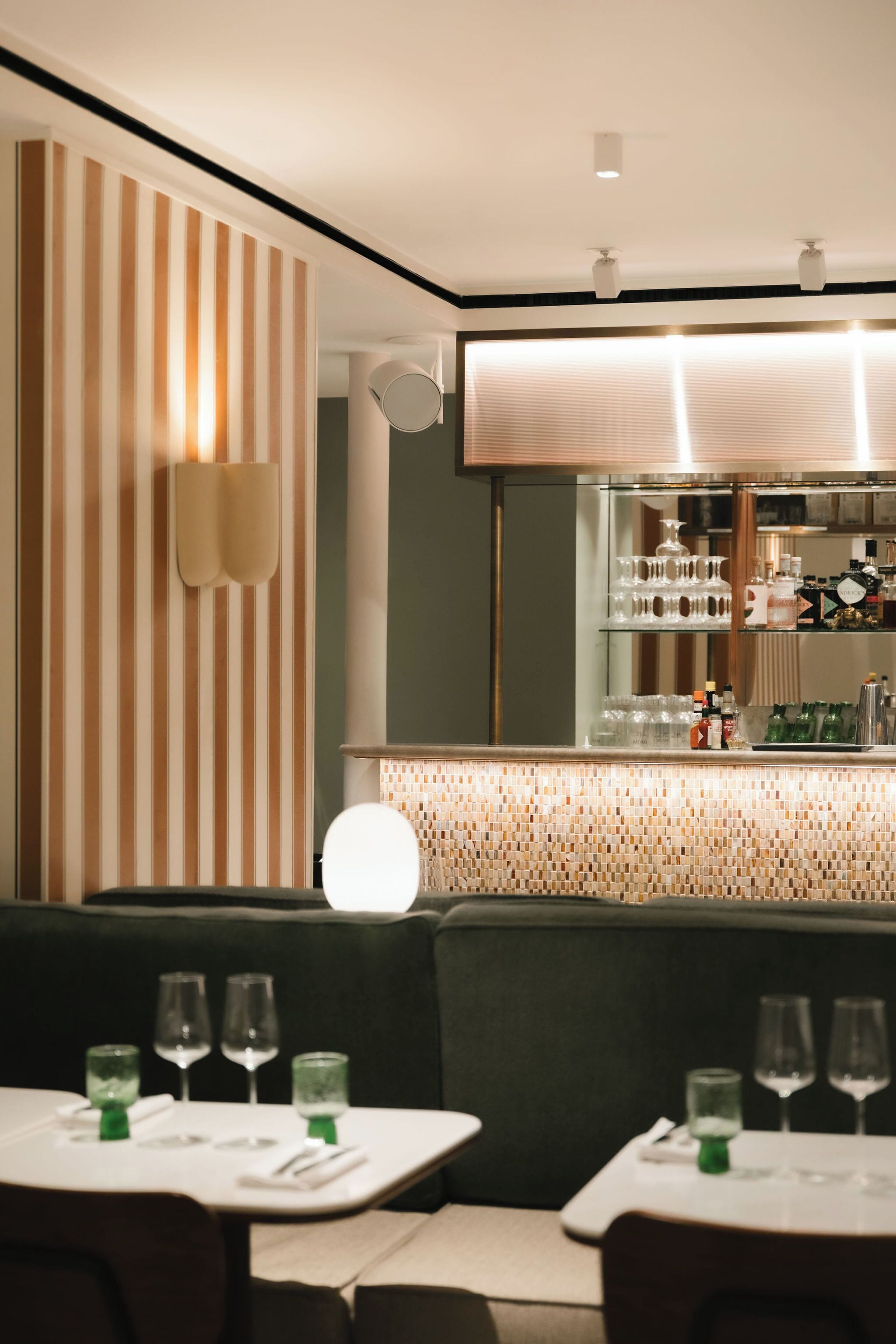 A restaurant with striped wallpaper, a bar area with shelves of glasses and bottles, and tables set with glasses and cutlery.
