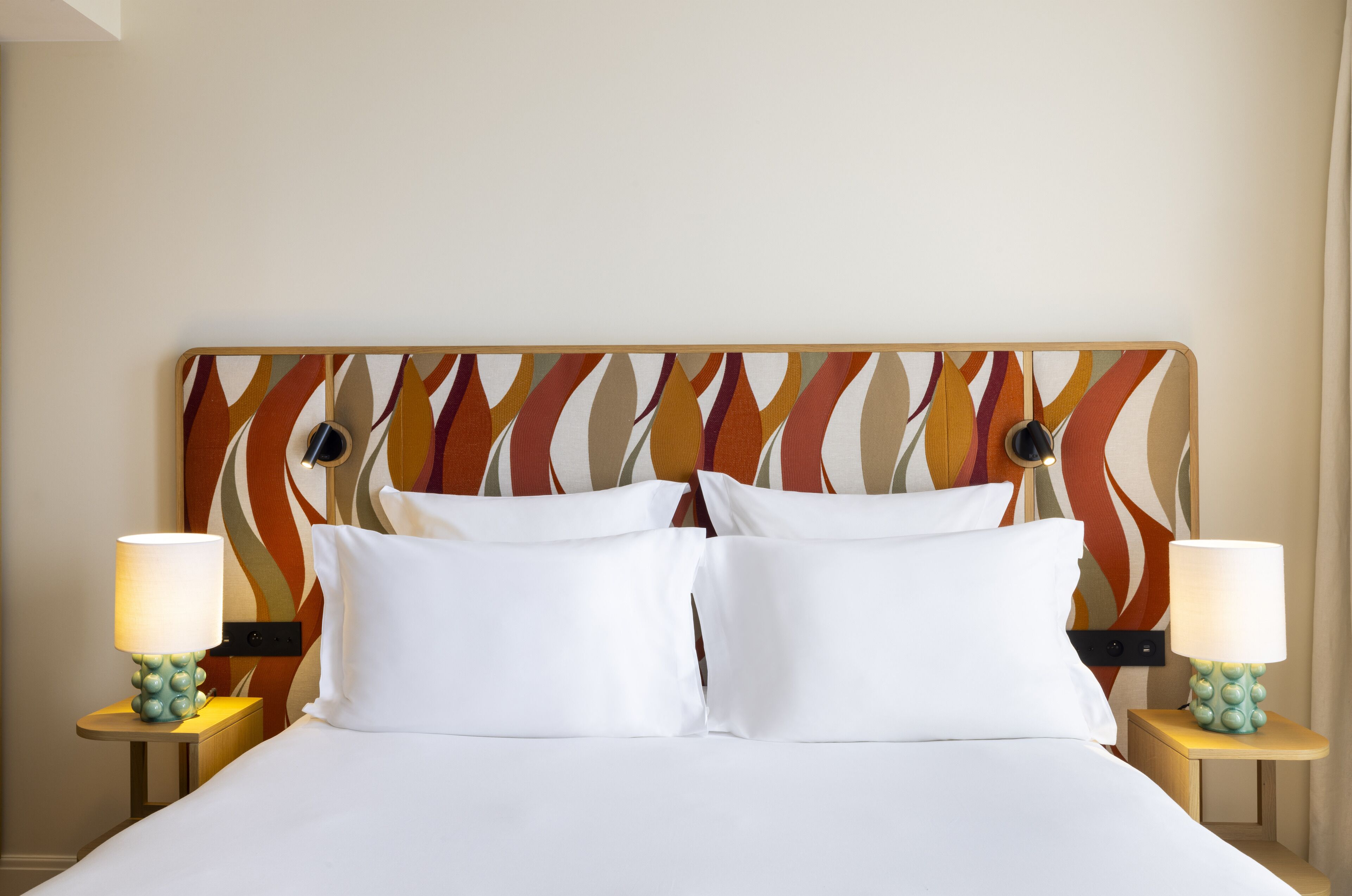 A hotel room with a large bed, two white pillows, a colorful headboard, and two bedside tables with lamps.