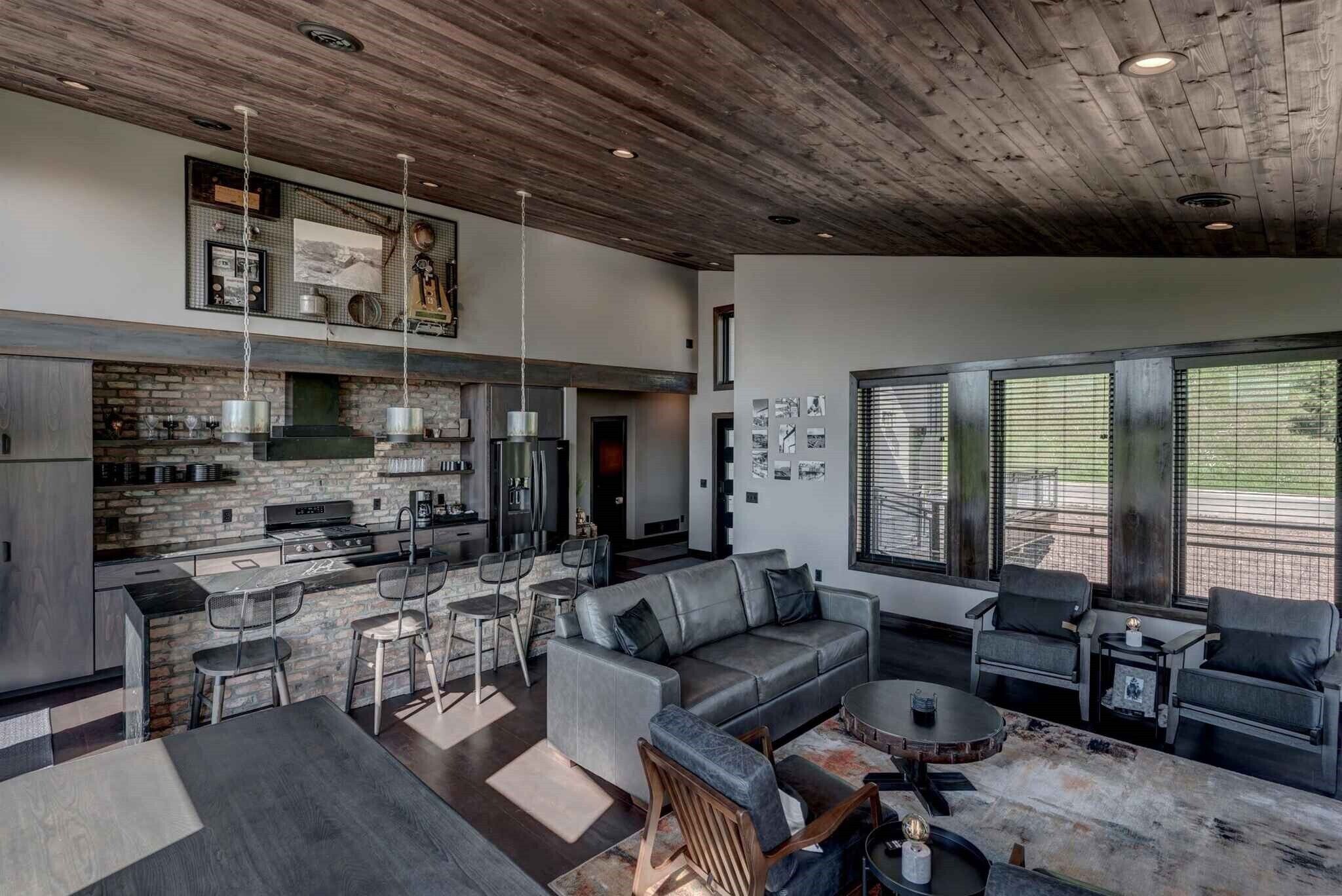 A modern kitchen with a stone backsplash, a dining area with a gray sofa and chairs, and a large window with blinds.