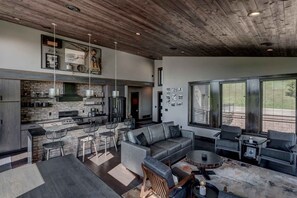 A modern kitchen with a stone backsplash, a dining area with a gray sofa and chairs, and a large window with blinds.