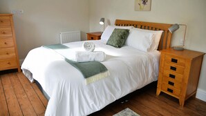 Een net opgemaakt bed met witte lakens, een groene plaid en een opgerolde handdoek. Er staat een houten nachtkastje met een lamp en een klein lade. De kamer heeft houten vloeren en een radiator onder het bed.