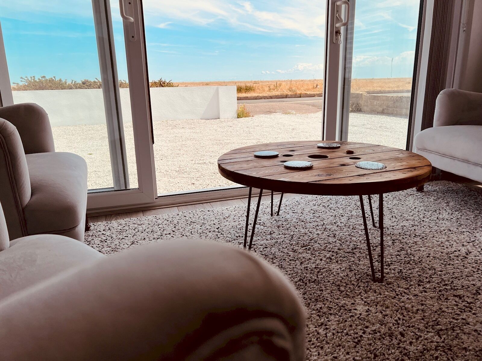 A modern living room with a round wooden coffee table, beige sofas, and large floor-to-ceiling windows offering a view of a desert landscape.