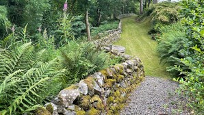 Una pared de piedra cubierta de musgo, un sendero de grava y abundante follaje verde.
