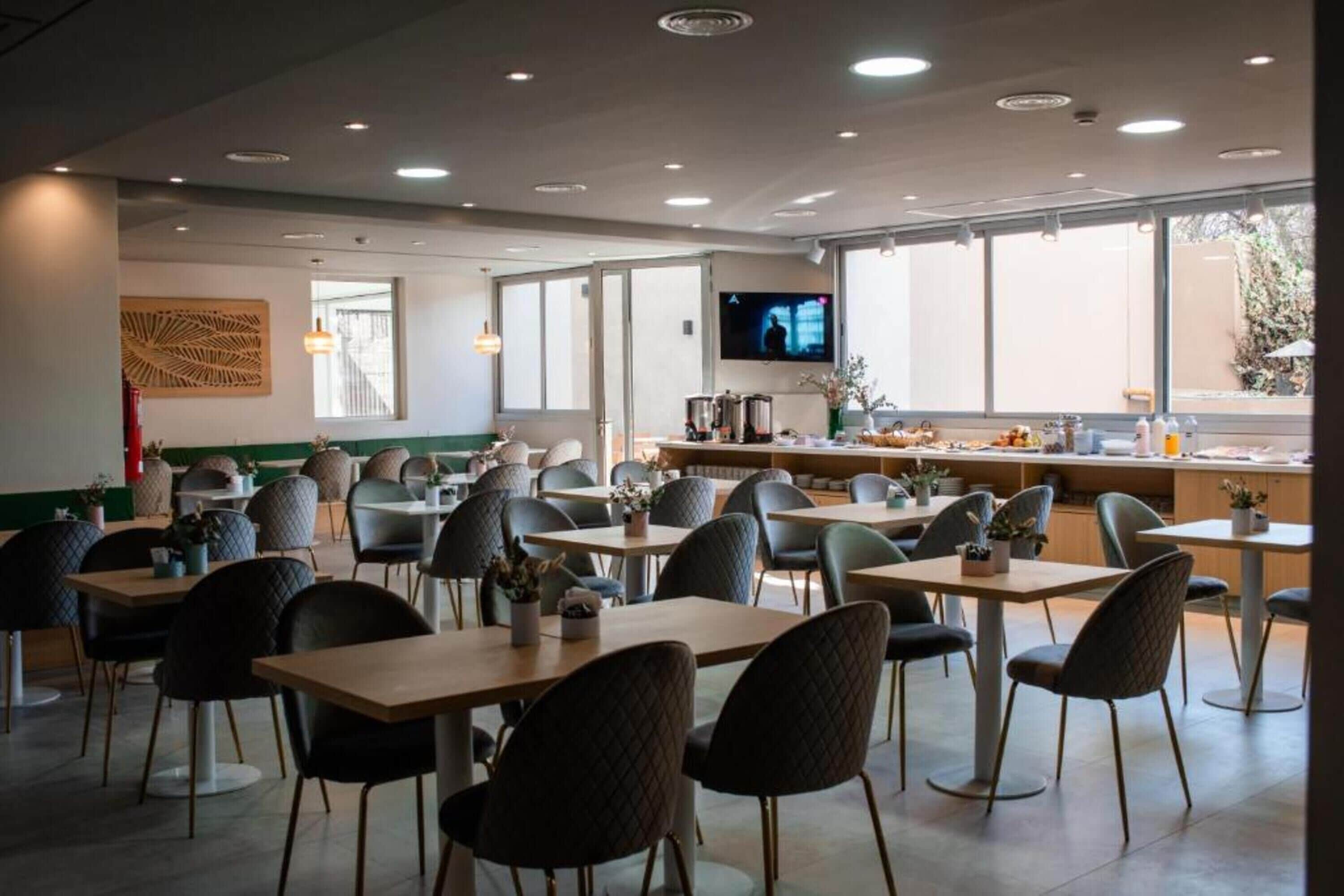 Una cafetería moderna con varias mesas y sillas, una pantalla de televisión y ventanas amplias.