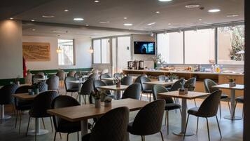 Una cafetería moderna con varias mesas y sillas, una pantalla de televisión y ventanas amplias.