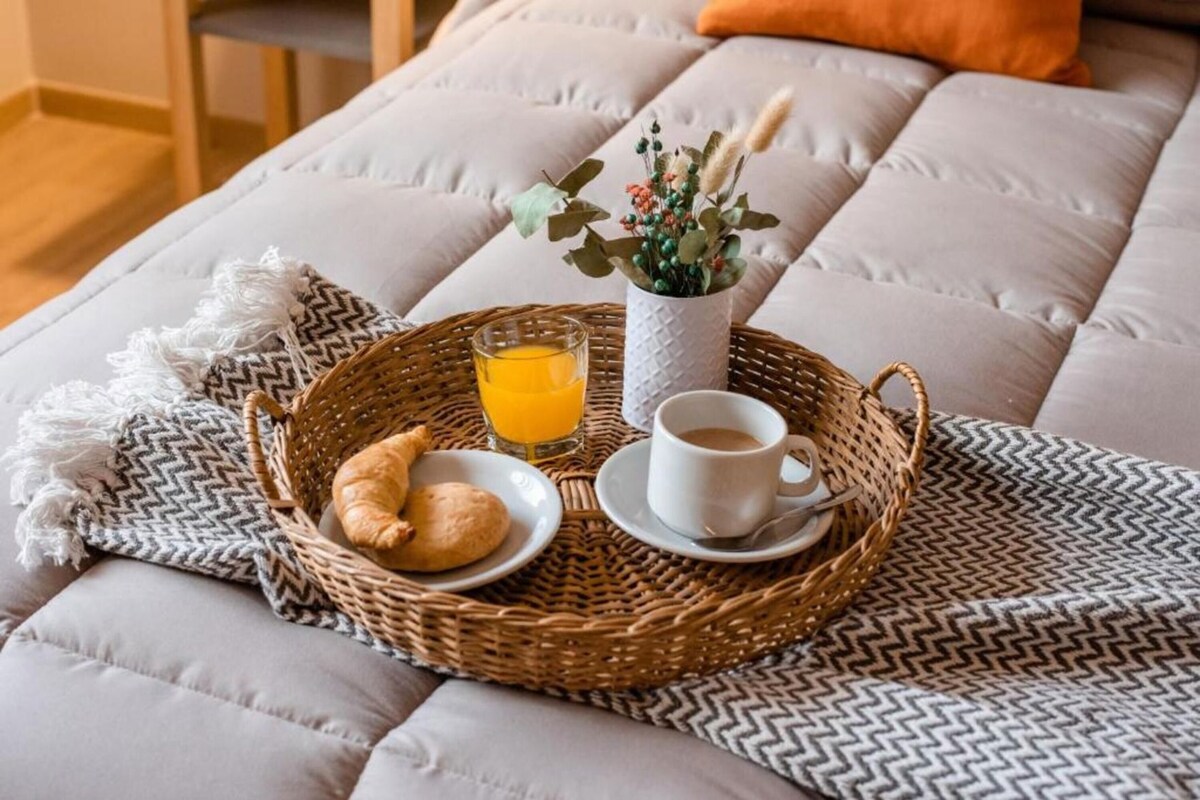 A breakfast setup on a bed with a basket containing croissants, a cup of coffee, a glass of orange juice, and a vase with flowers.