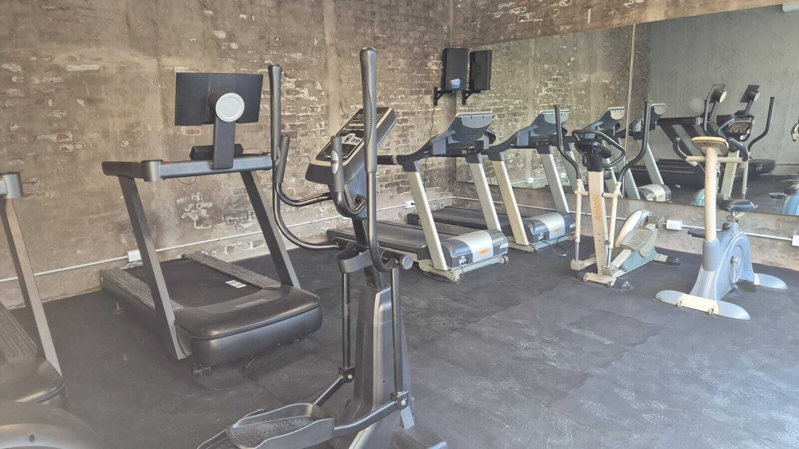 Una hilera de bicicletas estáticas y cintas de correr en un gimnasio con paredes de ladrillo y suelo de hormigón.