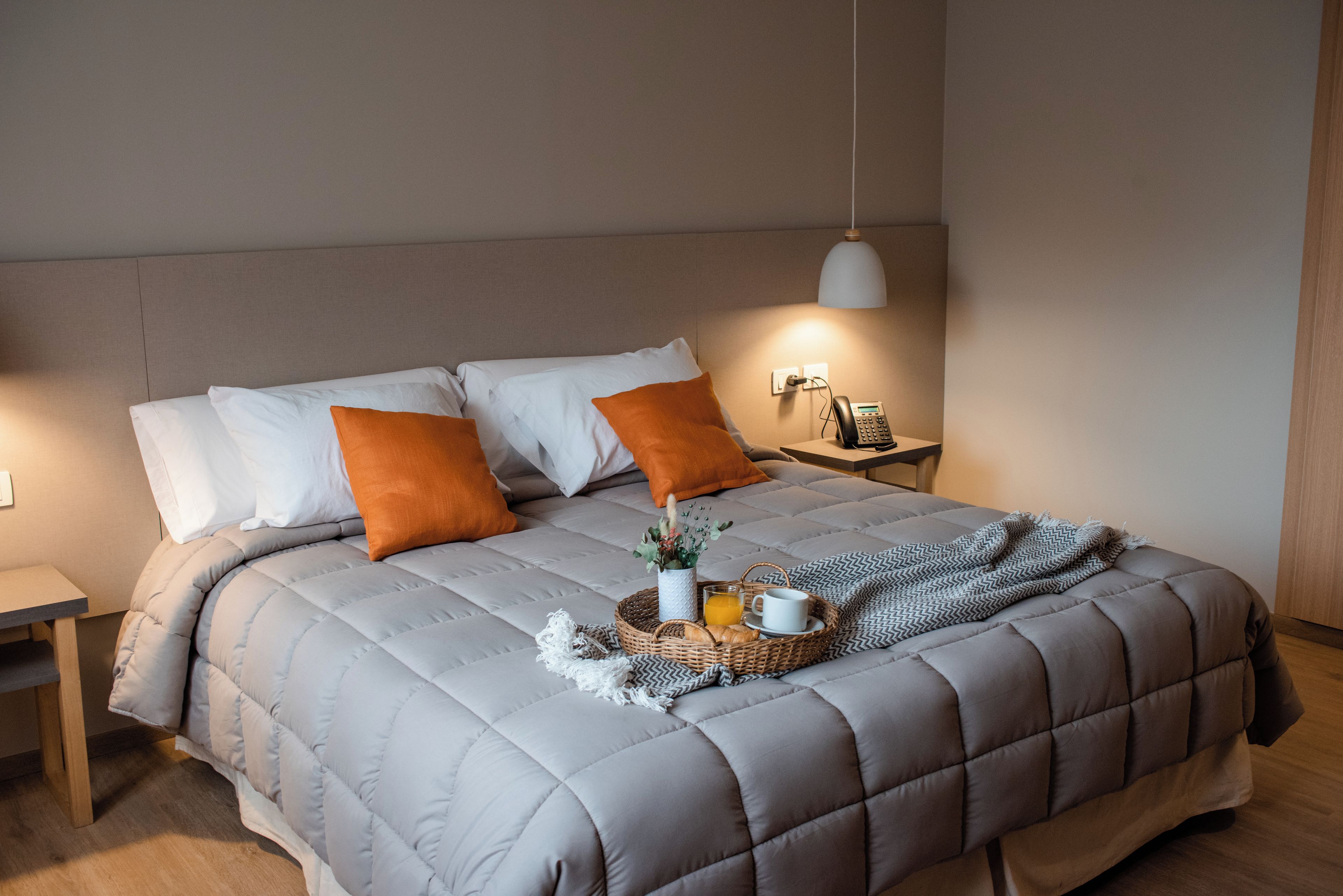 Una cama con un edredón gris, dos almohadas blancas y dos almohadas naranjas. Hay una mesita de noche con una lámpara, un teléfono y una cesta con una bebida y una taza.