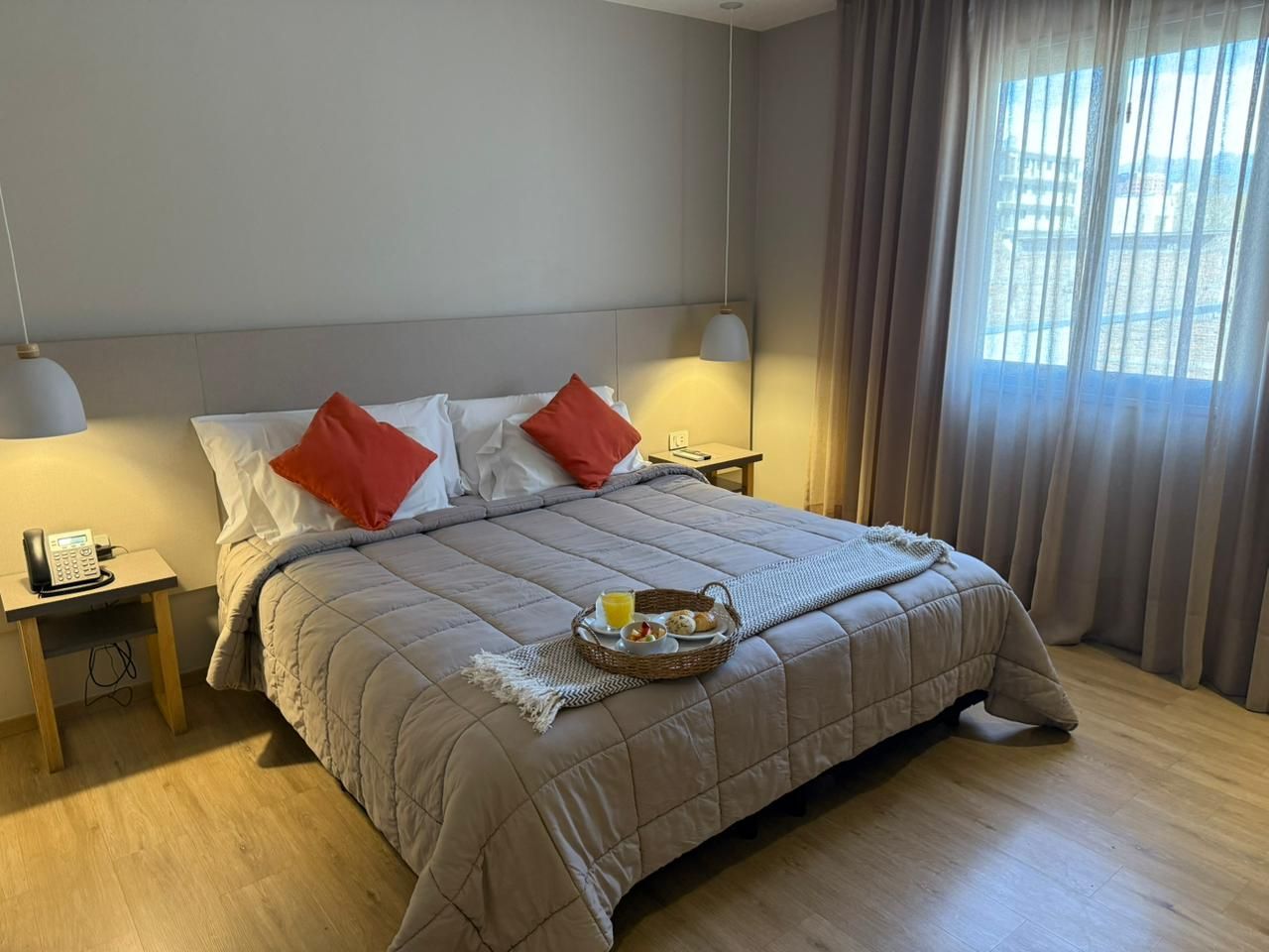 A hotel room with a bed, a bedside table with a phone, a tray with breakfast, and a window with curtains.