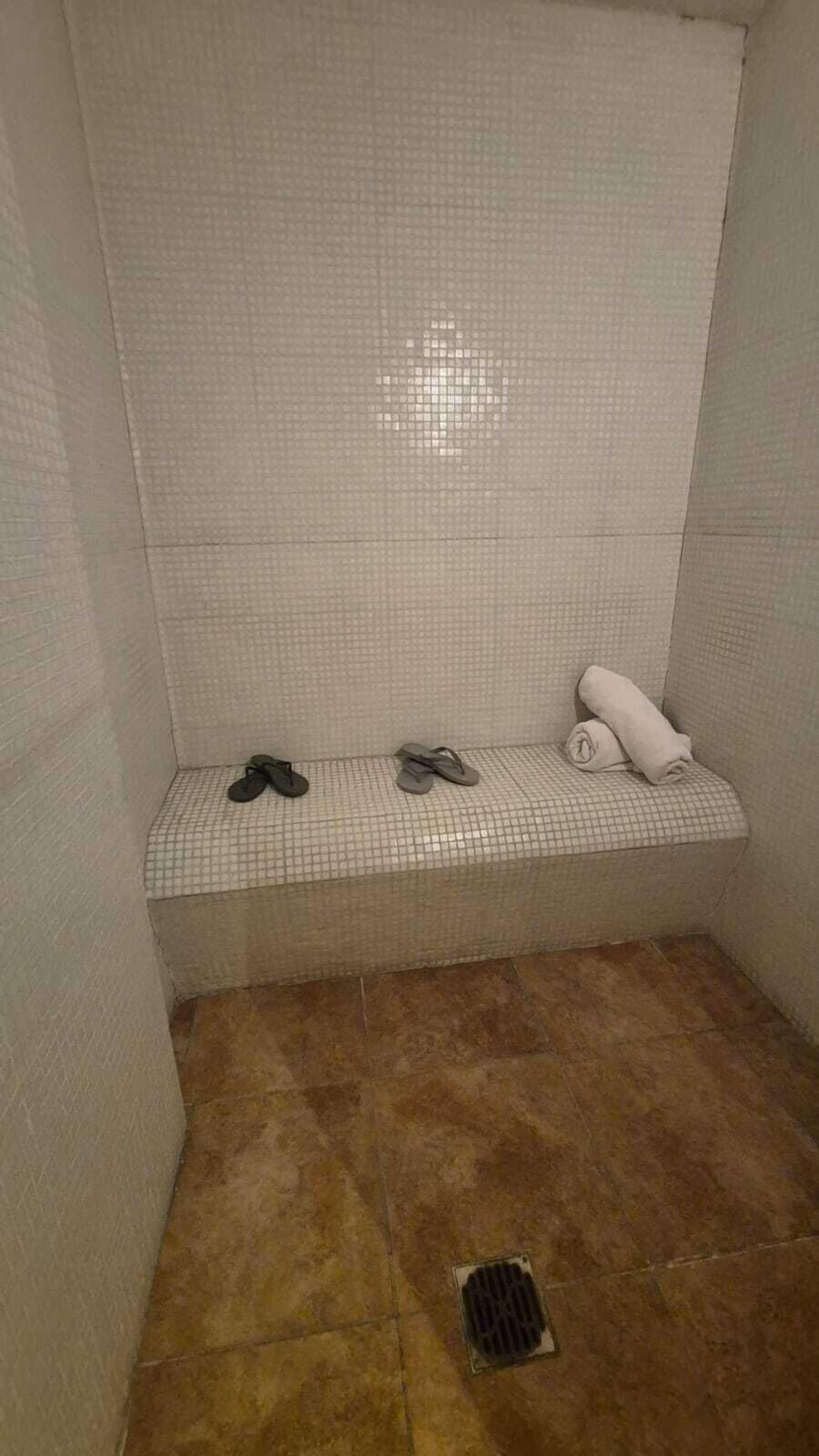 A small, tiled shower area with a bench, two pairs of flip-flops, and a rolled-up towel.