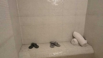 A small, tiled shower area with a bench, two pairs of flip-flops, and a rolled-up towel.