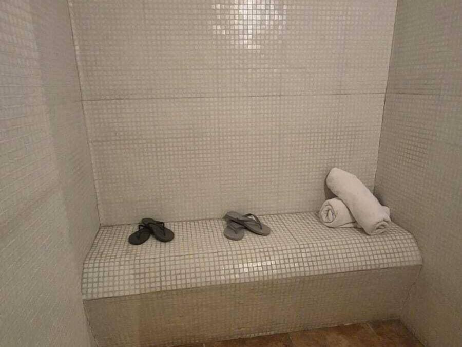 A small, tiled shower area with a bench, two pairs of flip-flops, and a rolled-up towel.