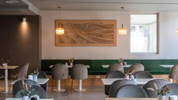 A modern restaurant interior with round tables and chairs, a green cushioned bench, and a decorative wooden panel on the wall.