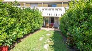 A courtyard with a pathway leading to a seating area, surrounded by lush greenery and a building with a balcony. - La Dunette - Two Bedroom Apartment, Sleeps 4 (La Grande-Motte)