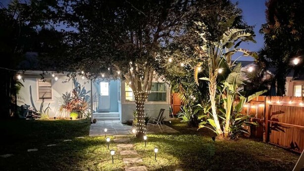 Un chemin de jardin bien éclairé qui mène à une maison avec une porte bleue, entourée d’une végétation luxuriante et de guirlandes lumineuses décoratives.