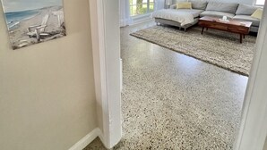 Un salon spacieux avec une grande fenêtre, un canapé blanc et une table basse. Deux tableaux représentant des paysages côtiers sont accrochés au mur.