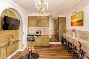 A modern kitchen with a built-in oven, a dining area with a long table and bar stools, and a TV mounted on the wall. - Merveil - Luxury Suite - Louvre - Rousseau (Paris)