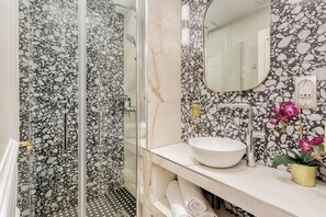 A bathroom with a glass shower enclosure, a white sink, and a marble-patterned wall. - Merveil - Luxury Suite - Louvre - Rousseau (Paris)