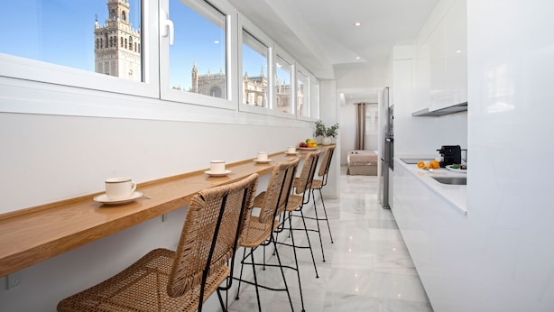 A modern kitchen with a long wooden counter, wicker chairs, and a view of a historic tower through large windows. - Genteel Home Constitución IV (Seville)