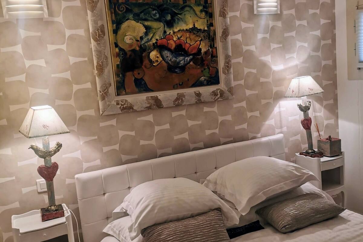 Ein Schlafzimmer mit einem weißen Bett, zwei Nachttischen mit Lampen, einem Bild über dem Bett und einer Wand mit Muster.