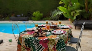 An outdoor dining table with a tropical tablecloth, surrounded by chairs, set against a poolside backdrop with lush greenery. - Blue Dream Cannes Guesthouse (Cannes)