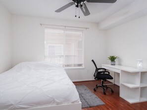Une chambre à coucher comprenant un lit, un bureau, une chaise, une plante en pot et un ventilateur de plafond.