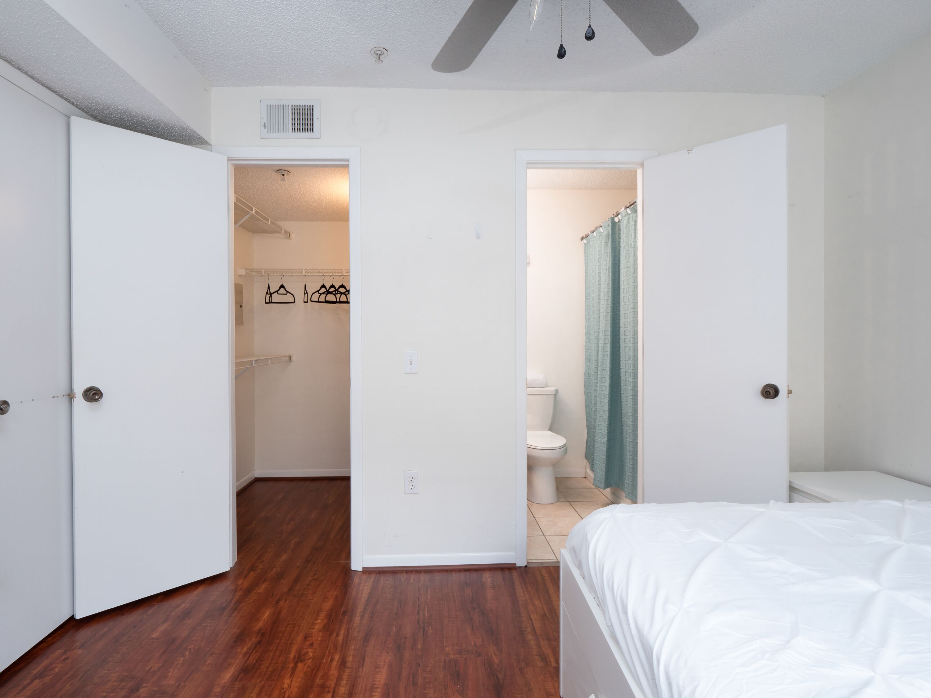 Un dormitorio con piso de madera, cama blanca y baño con inodoro y cortina de ducha.