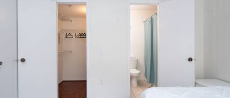 Un dormitorio con piso de madera, cama blanca y baño con inodoro y cortina de ducha.