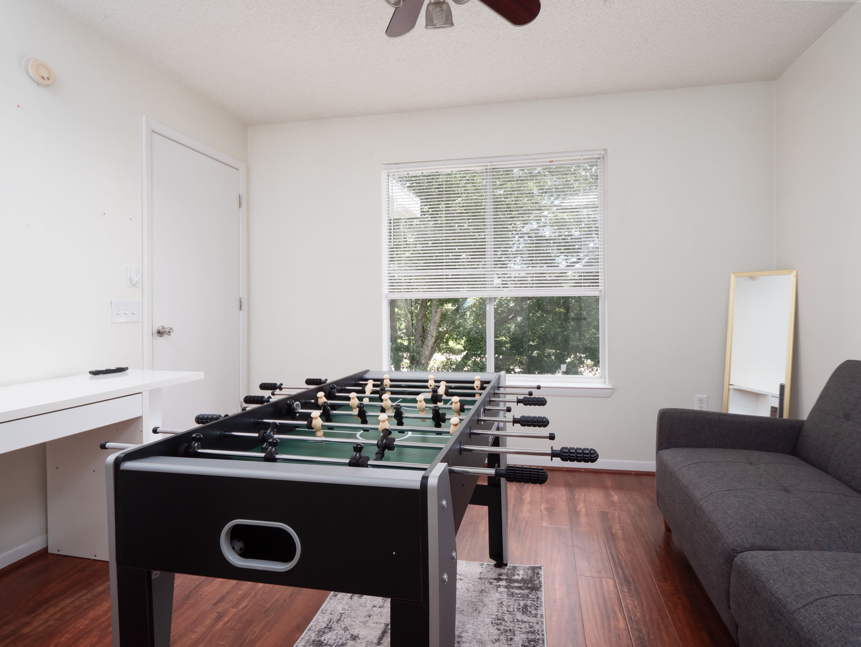 A room with a foosball table, a gray sofa, a window with blinds, and a white door.