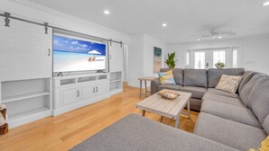 Uma sala de estar espaçosa com uma televisão de tela plana grande, um sofá modular cinza, uma mesa de centro de madeira e um rack branco para equipamentos eletrônicos.