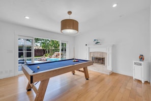 Une table de billard dans une pièce dotée d’un plancher en bois, d’une cheminée et donnant sur une cour arrière avec une piscine et des palmiers.