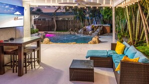 Uma área de pátio externa com piscina, assentos e uma televisão exibindo uma cena de praia.