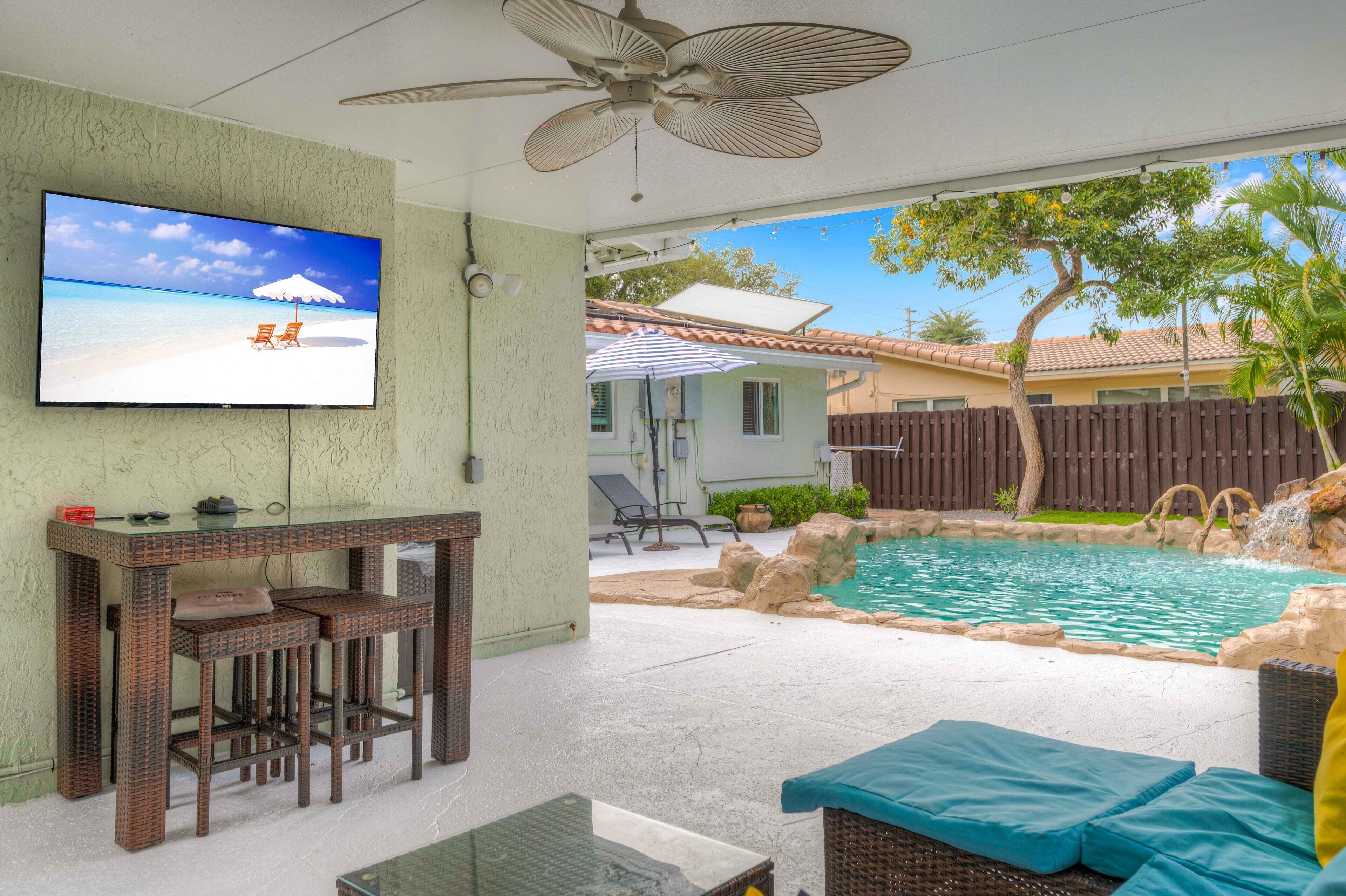 A pool area with a TV screen displaying a beach scene, a ceiling fan, and outdoor furniture.