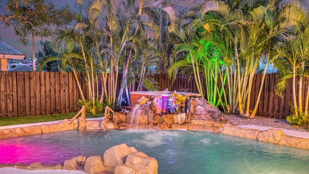 Piscina com cachoeira artificial, cercada por palmeiras e um muro de madeira.